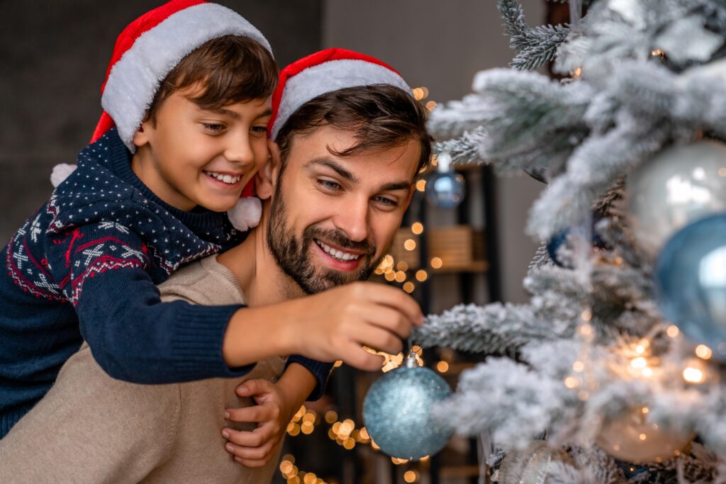 Smiling father and son putting ornaments and Christmas lights on tree during the holidays