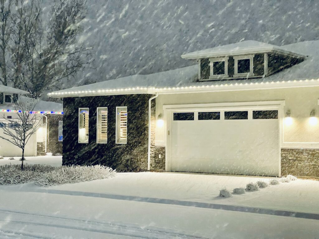 A home with holiday lights in a neighborhood during a snowstorm at night.