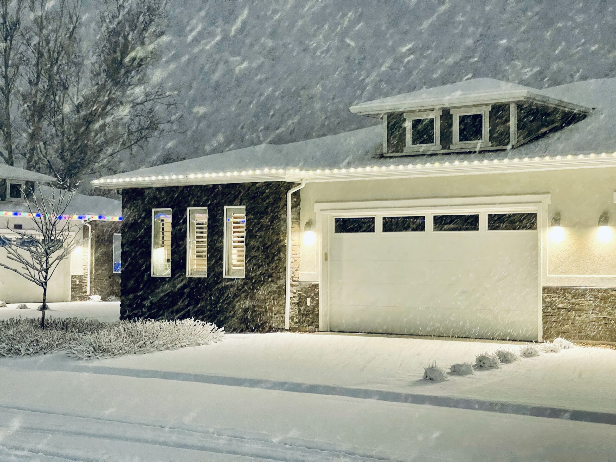 A home with holiday lights in a neighborhood during a snowstorm at night.